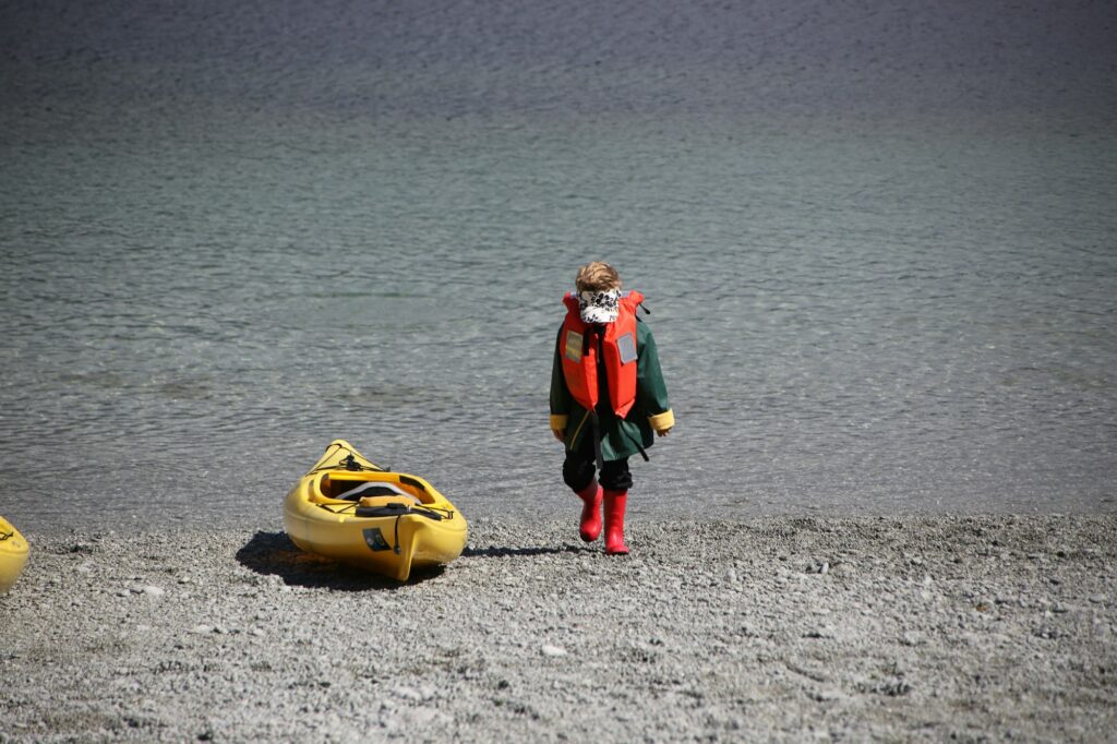 A small child in a red life jacket and rain boots walks along a pebble beach beside a yellow kayak
