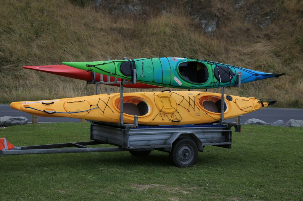Three kayaks strapped on a small two-wheel trailer parked on grass beside a road