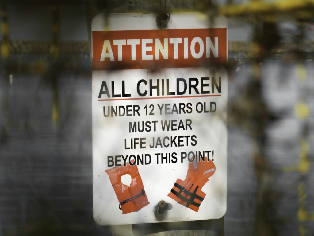 A posted sign at the water reading that all children under 12 years old must wear life jackets beyond this point