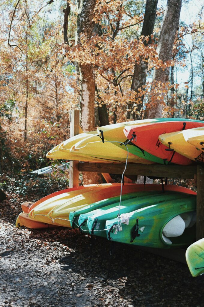 Sit-on-top kayaks stored outside on a two-tier wooden rack under autumn trees, off the ground, with a cable run through them
