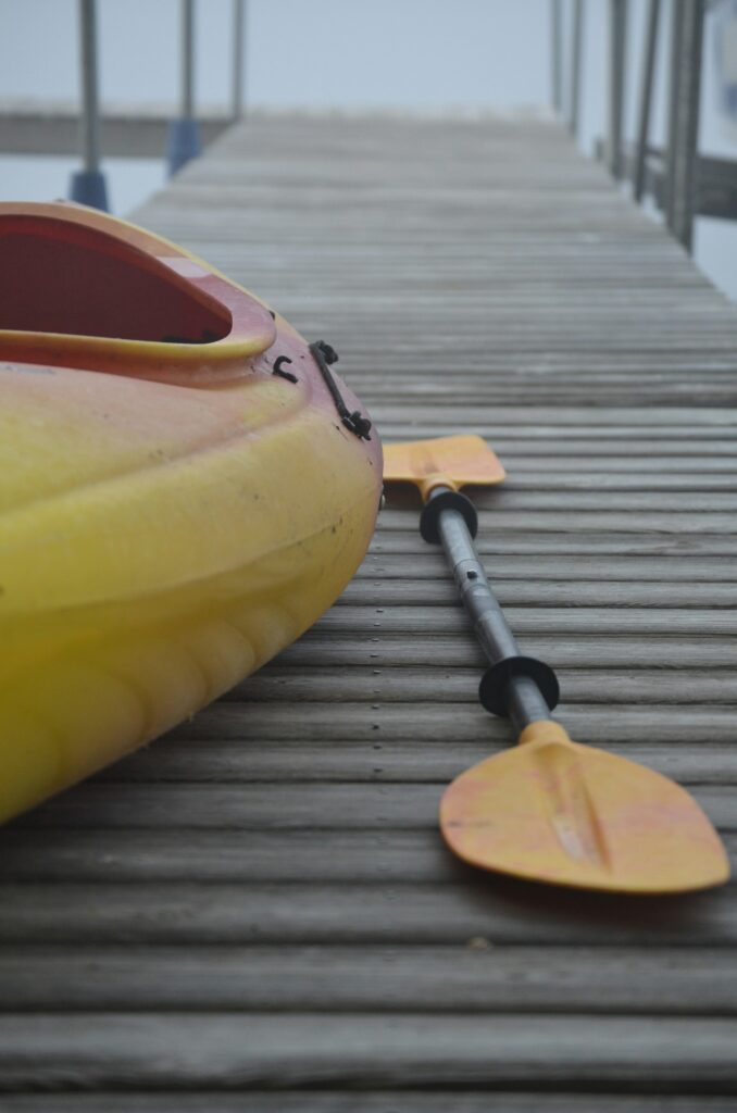 Close-up of the molded yellow and red plastic hull of a kayak resting on a wooden dock beside a paddle