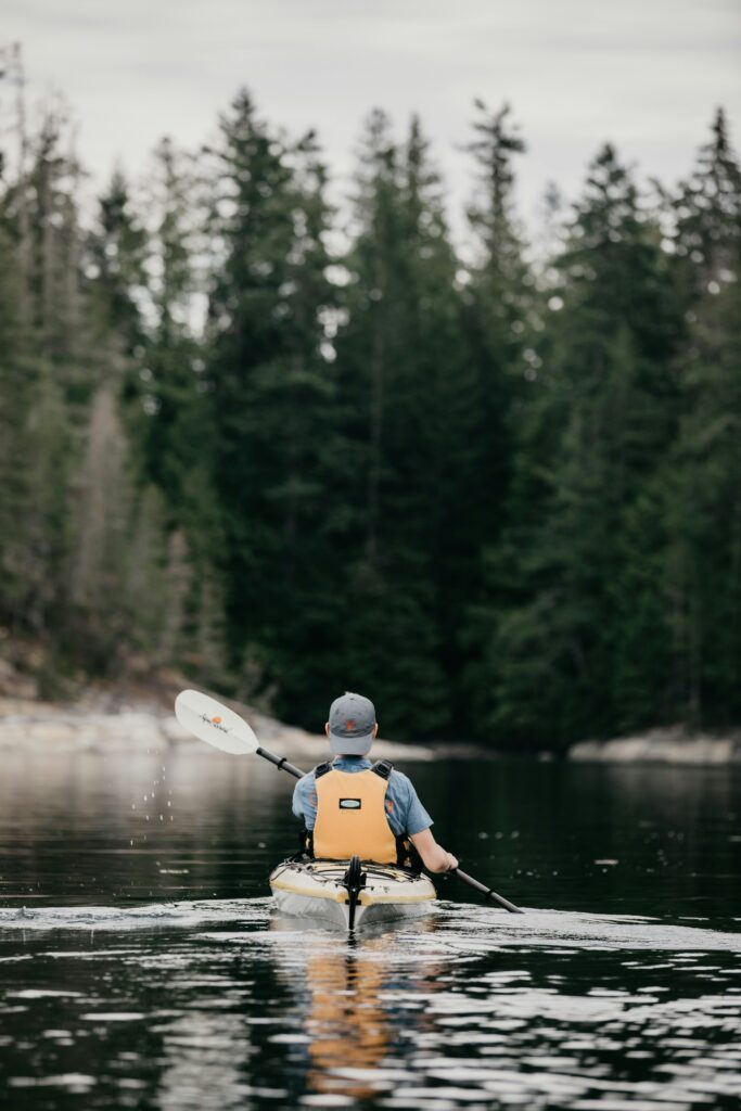 A paddler in a yellow life jacket heads across a calm, glassy lake toward a forested shore