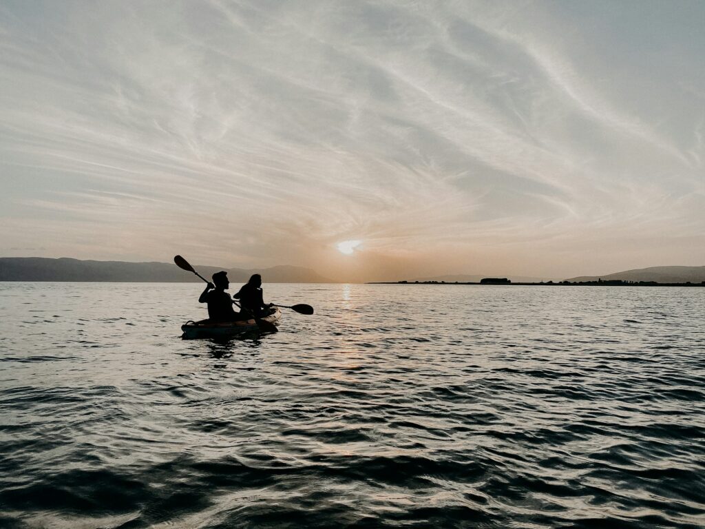 Two paddlers in one sit-on-top tandem kayak, silhouetted on open water at sunset