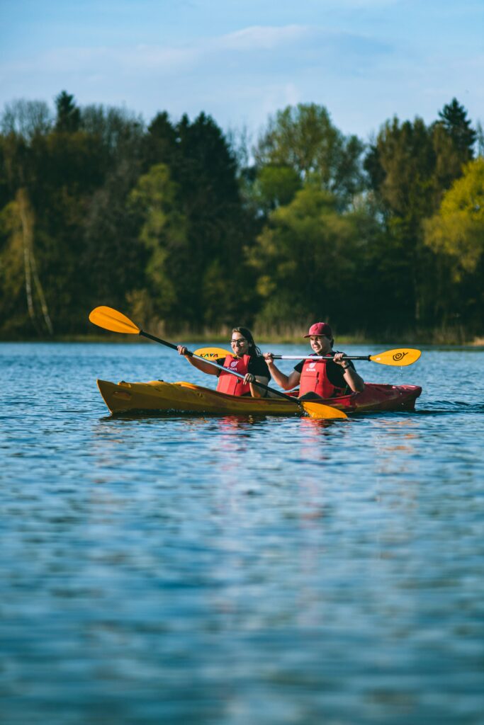 Two smiling paddlers in red life jackets share a tandem kayak on a calm lake lined with trees