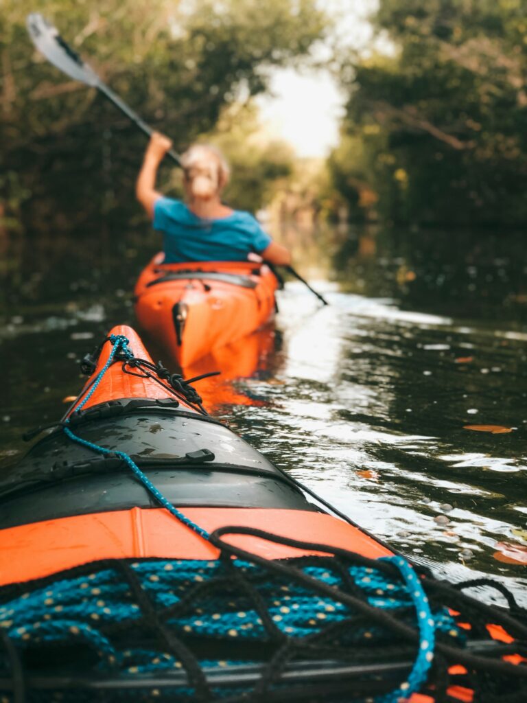 The loaded deck of an orange kayak with gear under bungee cords, another paddler ahead on a shaded river