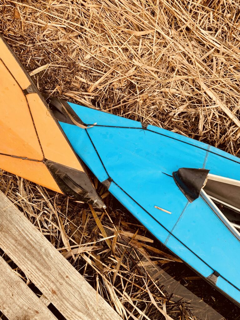The bows of two folding plastic kayaks, one orange and one blue, on dry straw beside a pallet
